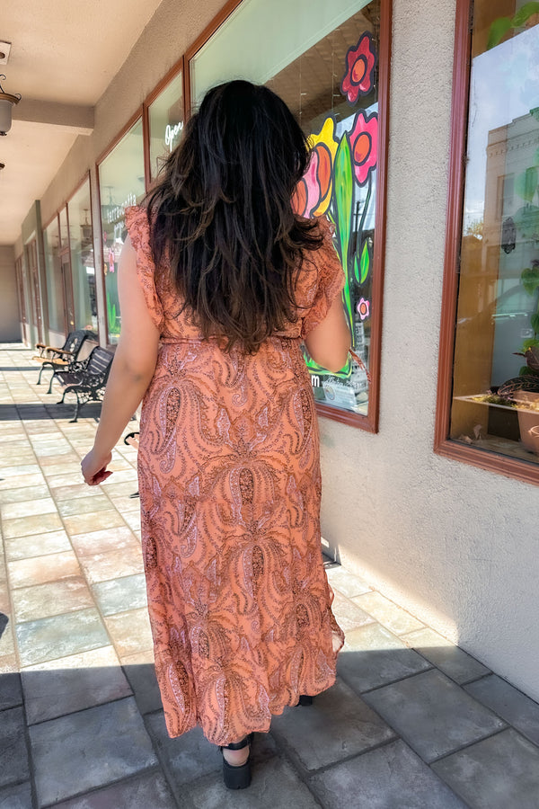 Back shot of a woman wearing a coral colored maxi dress with black sandals.