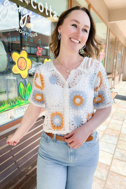 Woman wearing an off white, blue, and tan crochet button down top with light jeans.