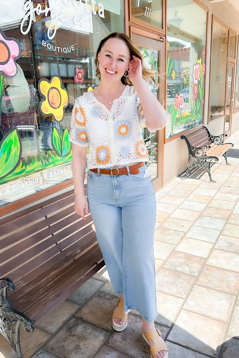 Woman wearing an off white, blue, and tan crochet button down top with light jeans and sandals.