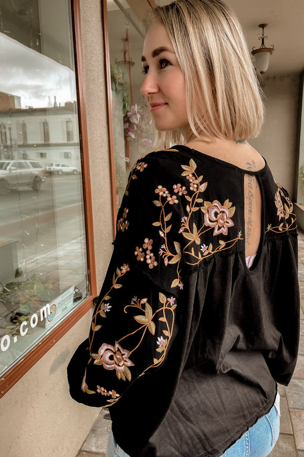 Blonde girl wearing a black embroidered drop sleeve shirt with light wash denim. (Back view)