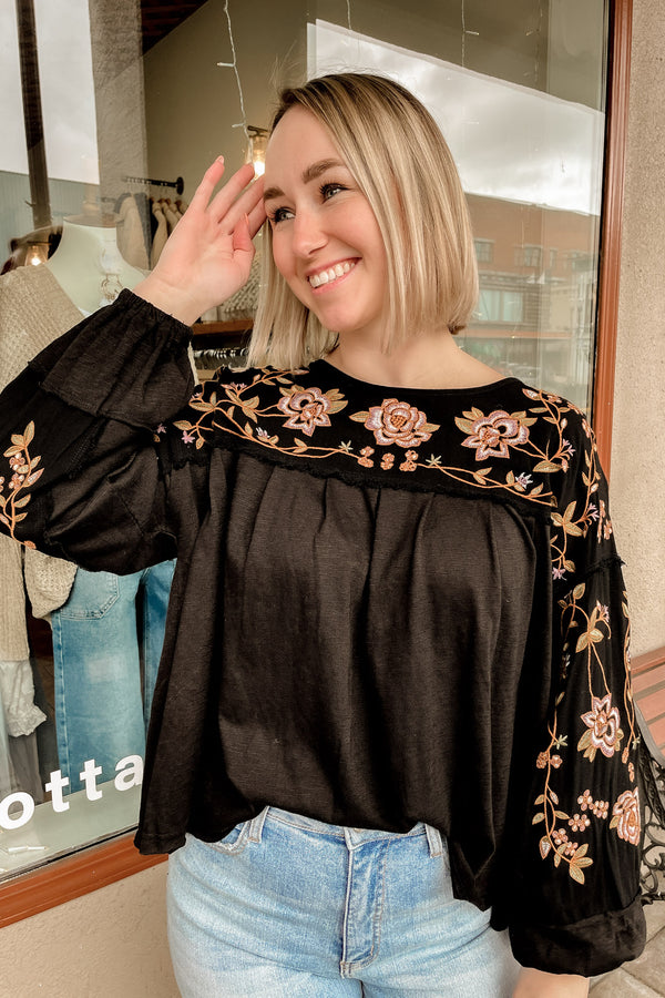 Blonde girl wearing a black embroidered drop sleeve shirt with light wash denim. 