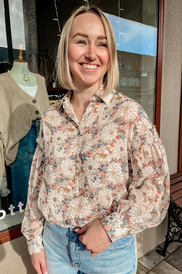 Blonde girl wearing a grey beige floral button down shirt with light wash jeans. 